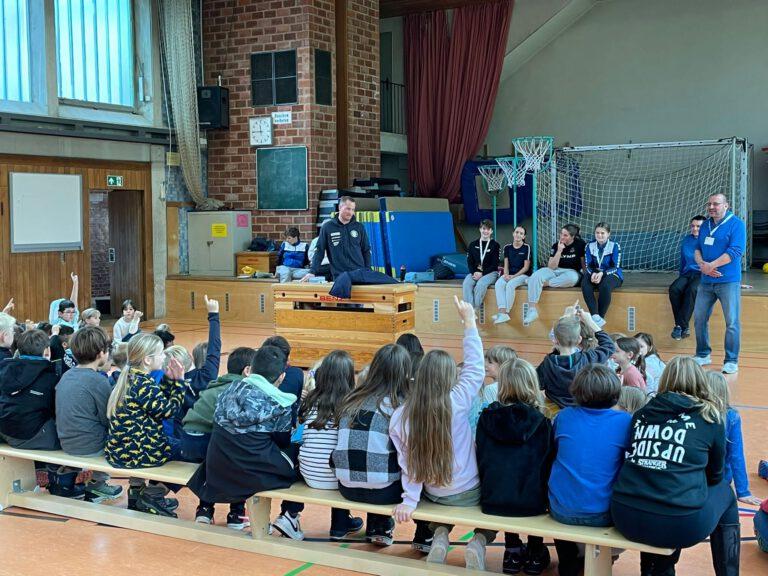 Kinder sitzen auf Turnbänken und hören einem Handballer zu